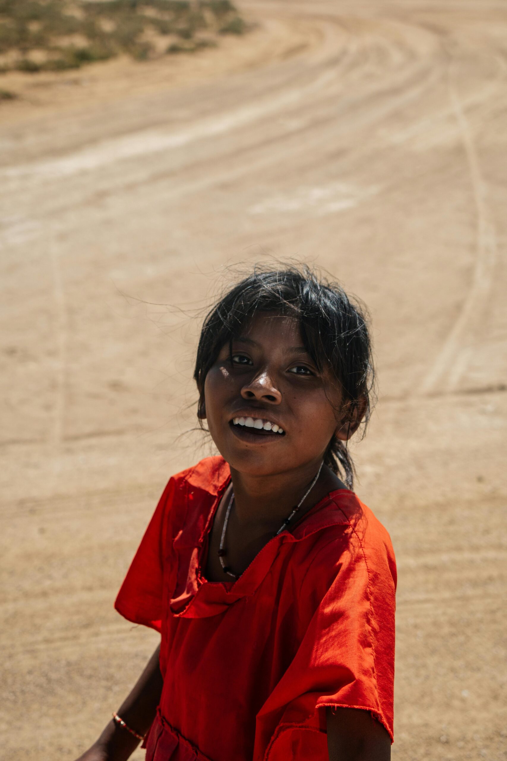 A cheerful child in a red dress stands on a sandy road in Guajira, Colombia.